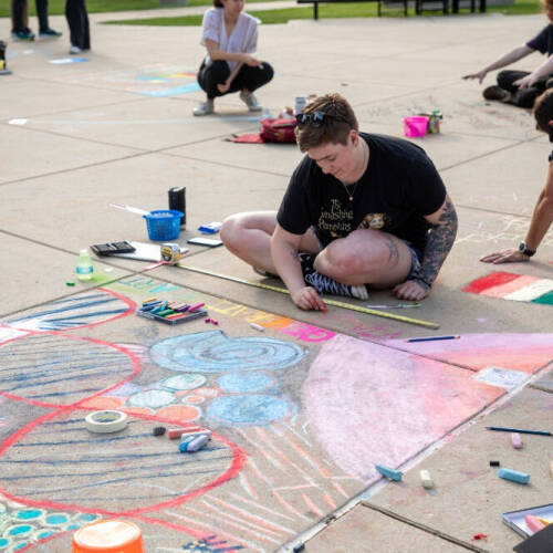 A person sits cross-legged on a concrete pavement using colored chalks to draw a large, geometric sidewalk painting in pastel shades, while other artists kneel in the background creating similar chalk artworks.
